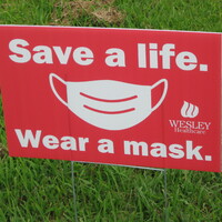 A red yard sign reading "Save a life. Wear a mask" with a graphic of a mask.
