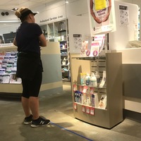 A woman in a pharmacy dressed in all black workout gear wearing a visor and is waiting in line. On the floor is a 6 feet piece of blue tape, measuring social distancing.