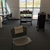 The corner of a library with windows. There are two book carts with books. There is a chair with a paper sign on it that says: Day 1 Quarantine.