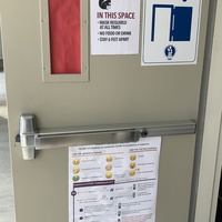 This is a picture of a classroom door that has several signs attached to it. The first one reads "In this space: Mask required at all times, no food or drink, stay 6 feet apart." The second message reads "Enter only" followed by a diagram of a door swinging open. A third message on the bottom of the door appears to depict a chart which showcases various information about COVID-19 symptoms.