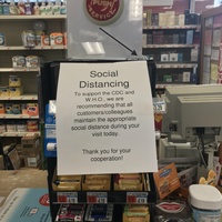 A register with a sign on it that says: Please press the button for a cashier and has an arrow pointing to the button. Below the sign is a paper sign that says: Social Distancing to support the CDC and W.H.O., we are recommending that all customers/colleagues maintain the appropriate social distance during your visit today. Thank you for your cooperation!