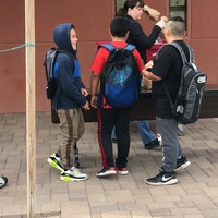 Three kids wearing backpacks are standing in front of a table outside. The kids are talking to one another. Behind the table is a woman who is putting paper lunch bags onto the table.