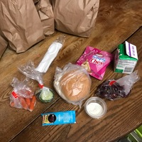 On a wooden table is a sandwich, slices of cucumbers, baby carrots, grapes, mayo, chex mix, and small cartoon of milk.