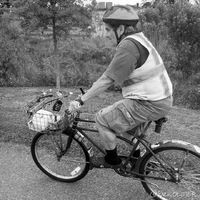 A man is riding a bicycle that is wearing a safety vest and bike helmet, on the front of the bike is a basket holding various items.
