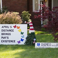 Photo of someone's front yard featuring two gnomes and a sign that reads "April's distance brings May's existence".