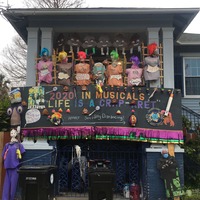 This is a picture taken of a person's house which is decorated with various items related to the COVID-19 Pandemic. A line of poop shaped emojis with smiley faces hang from the roof, under which sits a line of dummies with skulls for heads each labeled under a different spelling of the name "Karen", with a sign underneath stating that each dummy did not wear their mask. A banner underneath these objects reads "2020 in musicals: 'Life is a Crap-aret'". The bottom of the banner encourages the reader to follow social distance protocols. Several mannequins dressed in medial gear or face masks flank the banner.