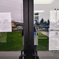 Two doors in front of a store have multiple papers taped on the inside of the glass informing patrons of their changed store hours and the decrease in people in the lobby.